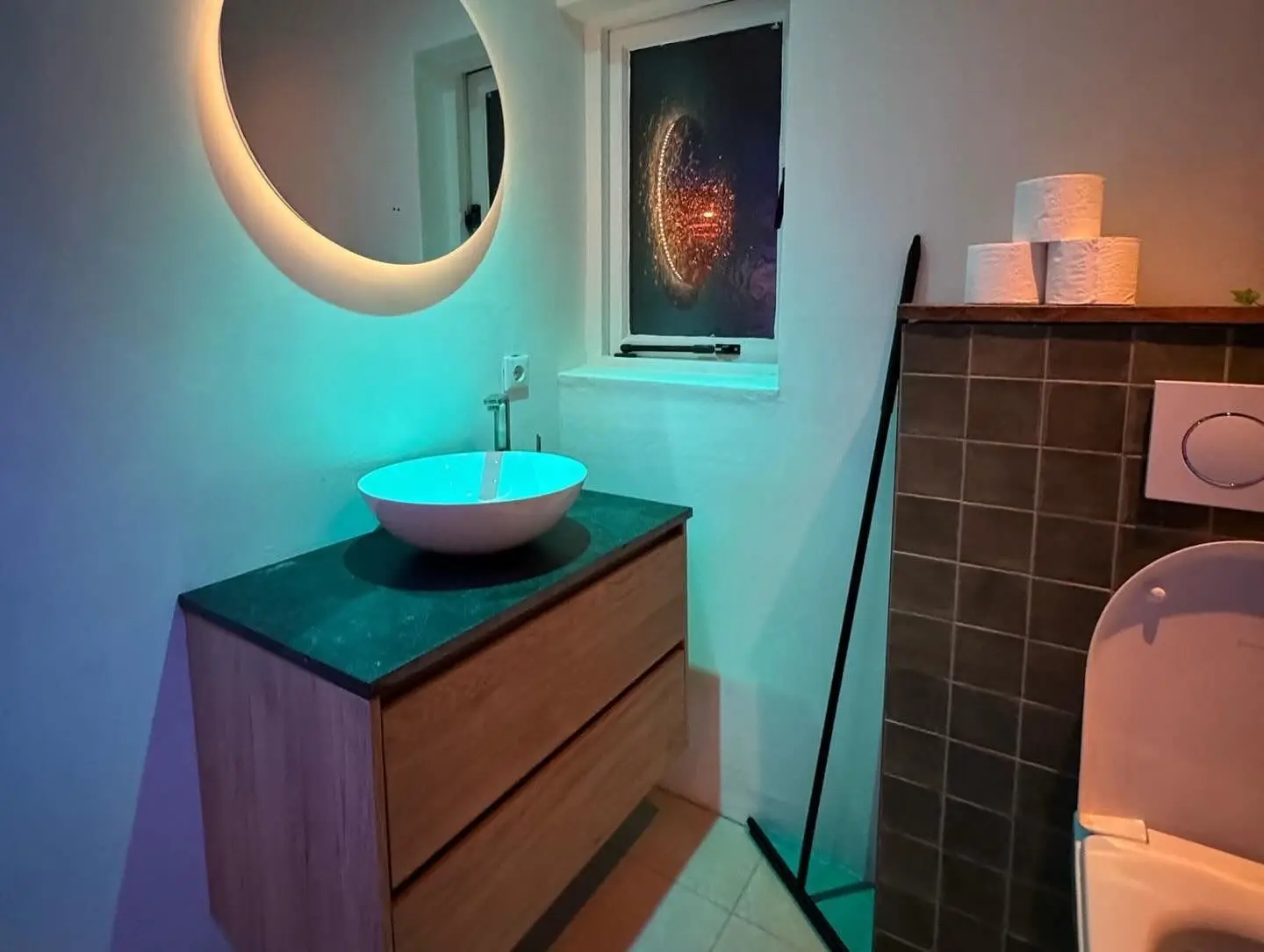 Bathroom vanity with a round illuminated mirror and a ceramic sink under colorful lighting.